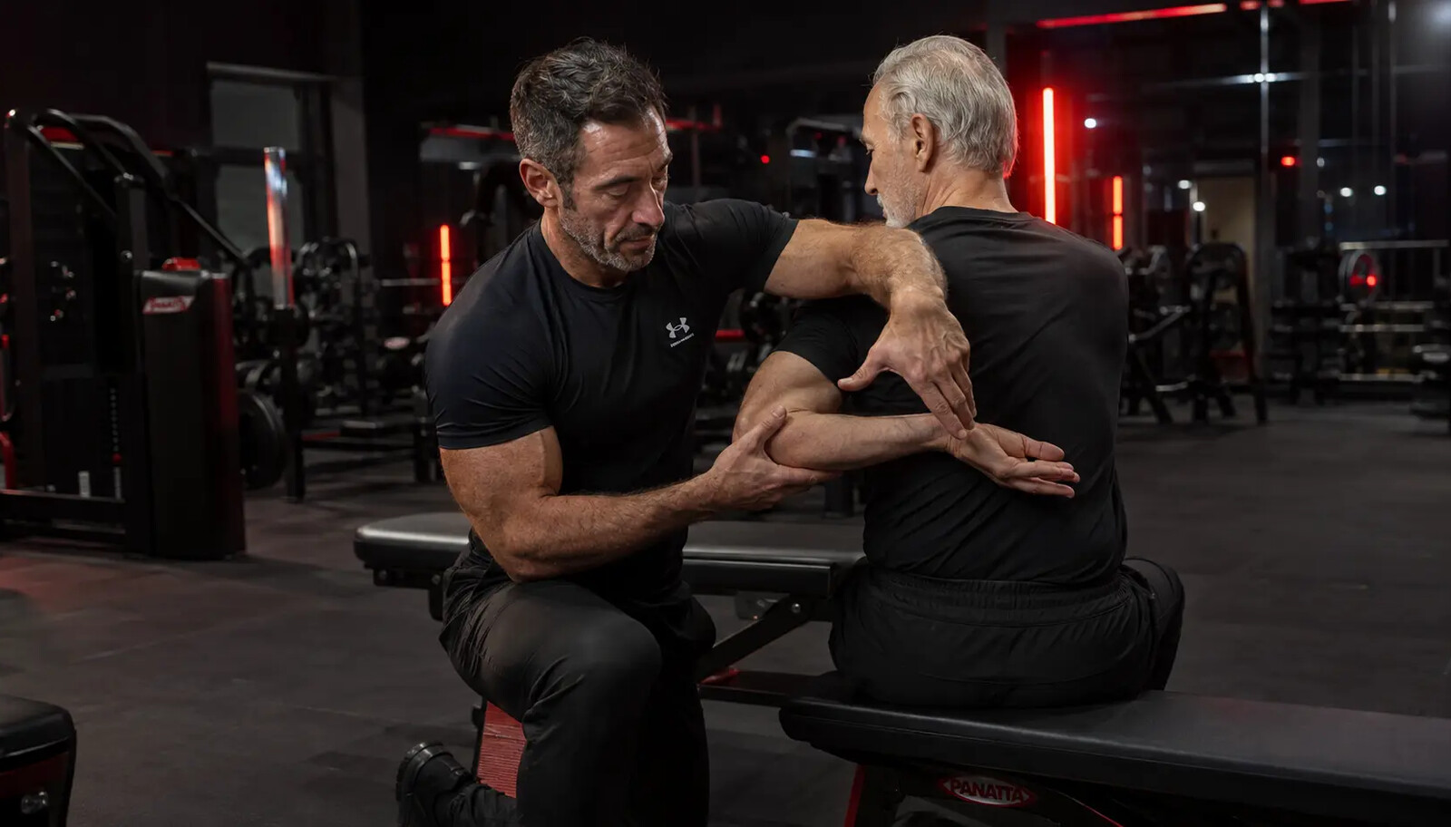 A fit, athletic man in his late forties sitting calmly on a bench between sets in a professional gym, towel over his shoulder