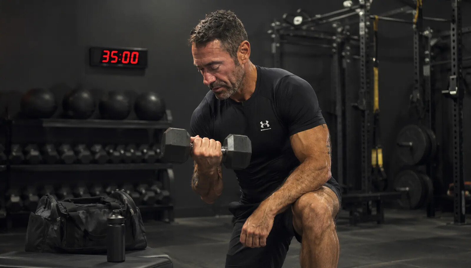 A fit, healthy adult in their forties performing a focused dumbbell lunge in a dark professional gym